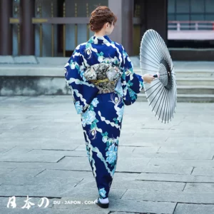 Femme en kimono fleuri avec ombrelle, marchant paisiblement sur un chemin de pierre.