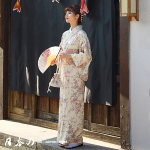 Femme en kimono pastel élégant, tenant un éventail, devant un fond rustique traditionnel.